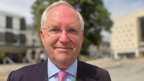 Chris Nelson - an older man in a suit with grey hair and thin rimmed glasses - smiles as he stands in a city centre square.