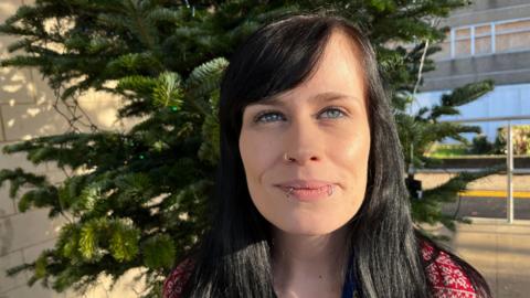 Gemma, a woman, with black hair, smiling for the camera. The tree is behind her. She is outside and there is a 1960s concrete building in the background.