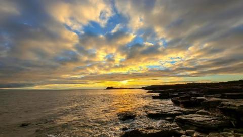 The sun sets on the horizon creating yellow reflections arching up to the clouds and reflected in the sea below