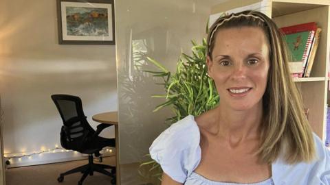 A woman with blonde hair looks at the camera, with a plant and office furniture behind her.
