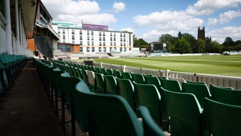 photograph taken from the stands of New Road showing green plastic seats in rows in front of the camera. A white building is pictured on the edge of the stadium with trees and Worcester Cathedral overlooking the pitch.