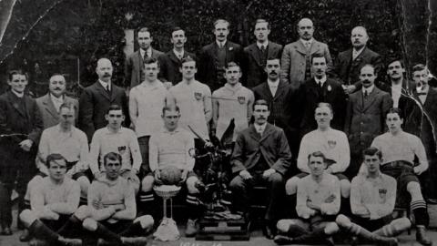 A black and white photograph from 1912 showing 27 men sitting and standing in rows. Many of the men are dressed in suits with waistcoats, but others are dressed in football kit, and one is holding a football. There is  large (2ft/0.6m) trophy showing a winged male figure holding a laurel leaf and trampling on a figure below