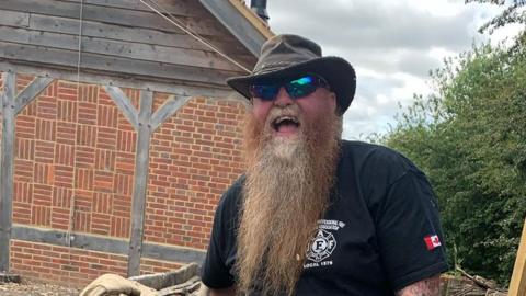 A man with a long beard, wearing a hat, holding a handmade chisel laughing