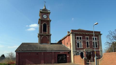 The Old Town Hall which is a red brick building with a clock tower on the left on top of a one storey extension. The main building on the right is a rectangular building with three storeys. It looks run down with the windows boarded up.