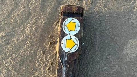 A waterlogged footpath signpost from Shropshire which has been broken off at the bottom and is lying in the sand