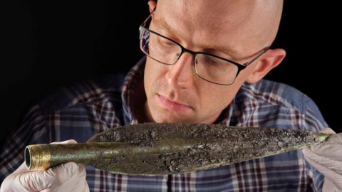 Dr Matthew Knight, who is a caucasian man wearing glasses, is holding and examining the spearhead found in the Carnoustie Hoard.