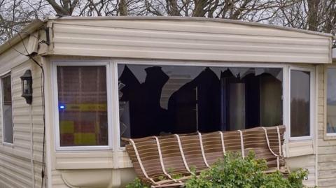 The outside of a badly damaged caravan. Its front window is smashed.