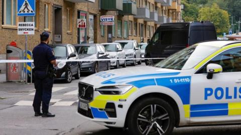 A police car stands outside a crime scene as a policeman in uniform looks on beyond a cordon in Sweden