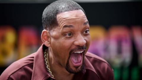 Will Smith is looking slightly to his right with an animated expression on his face: mouth wide open showing his tongue and upper teeth in a broad smile with his eyebrows high on his forehead. He has short cropped hair shaved into sharp edges, and is wearing an open necked burgundy shirt with broad lapels. He has several thick gold chains around his neck. 