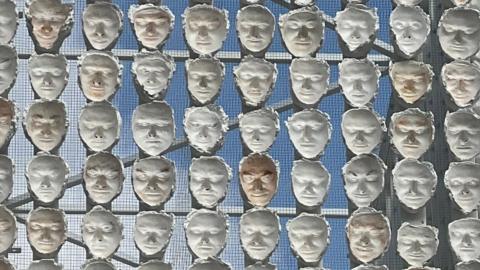 Plaster casts of the faces of transgender and non-binary people arranged in rows on a mesh background