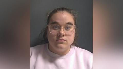 Zara Lancashire: a woman with pulled back brown hair wearing pale-rimmed glasses and a pink round-necked top.