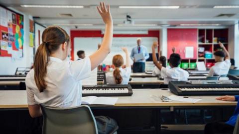 An image of a girl at the back of a classroom with her hand in the air. The image is taken from behind. 