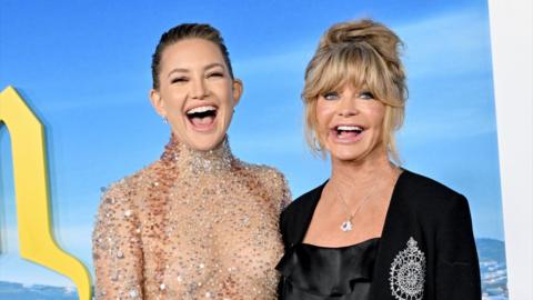 Kate Hudson and Goldie Hawn stand together with big smiles - Kate wears a sequin top, Goldie wears a black jacket