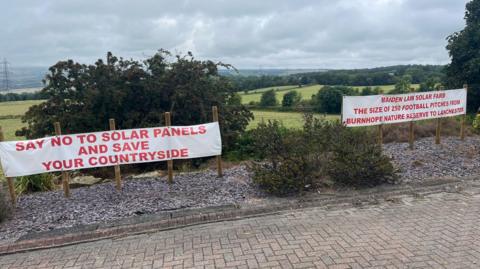 Two white banners with red letters attached to a fence on the edge of fields. One reads: "Say no to solar panels and save your countryside" and the other says: "Maiden law solar farm the size of 250 football pitches from Burnhope nature reserve to Lanchester".