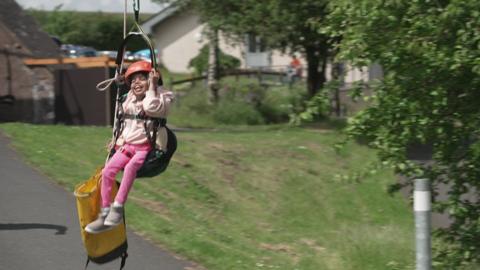a girl in a light pink hoodie and darker pink trousers on a zippier in a red helmet