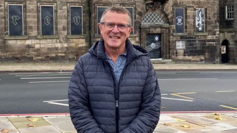 Jon Plant, a man wearing glasses, as well as a blue shirt and a dark-coloured jacket, is standing by the side of a road. He is smiling for the camera. There is a stone building in the background.