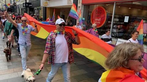 People walk down a street of shops holding up a giant long rainbow flag. They also wear colourful clothes and wave other smaller flags