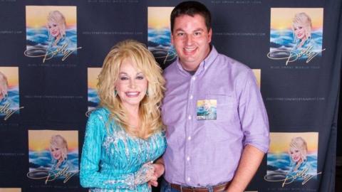 Dolly Parton with voluminous blonde hair, a sparkly blue and silver long-sleeved top is smiling next to a man who she is holding the hand of. The man has short brown hair and a purple long-sleeved top on. Behind them is a curtain full of repeated photos of Dolly and her signature.