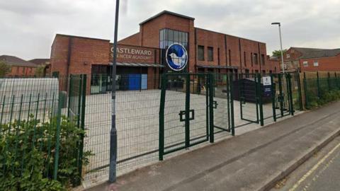 A red-brick school building behind a green fence with a grey sky behind