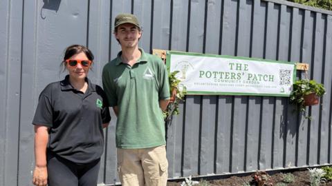 A woman wearing a black polo shirt and a man wearing a green polo shirt are standing next to a grey shipping container which has a white banner on with green lettering which promotes The Potters Patch community garden. There is a hanging basket on each end of the banner attached to a wooden plank.