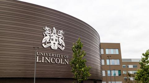 The exterior of a University of Lincoln building with a logo on the front. The sky is overcast.