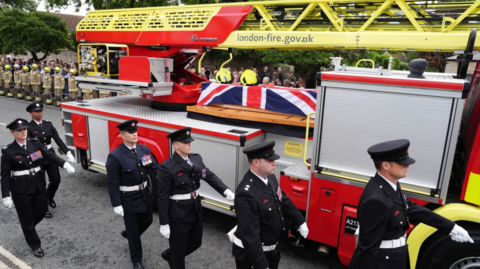 A turntable ladder carrying the coffin of Firefighter Martyn Sadler passes Bicester Fire Station, Oxfordshire, ahead of his funeral. The coffin in draped in the Union Flag and the vehicle is flacked by firefighters in full dress unifor