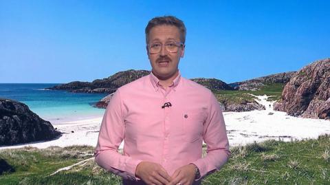 Weather presenter in a pink shirt with short hair and glasses, in front of a picture of a beach with white sands, rocky shores, machair and blue seas and sky.