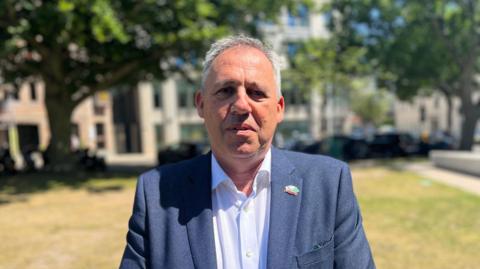 Infrastructure Minister, Constable Andy Jehan, outside the Jersey Government building on Union Street, wearing a white shirt and blue blazer