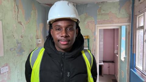 Takunda Magwaza standing in a room that has bare walls and is in a bad state of repair. He is wearing a hard-hat, black coat and high-viz jacket and is looking straight at the camera.