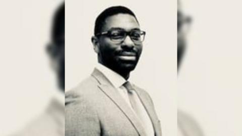 A sepia image of Damo Awobajo, a man wearing glasses, a suit jacket plus a shirt and tie.