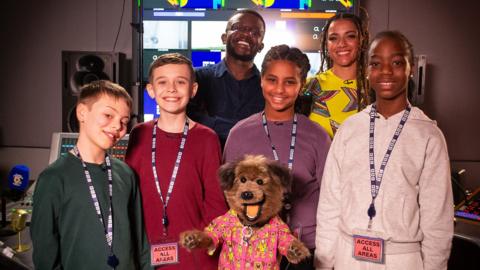 Newsround’s De-Graft Mensah, Saturday Mash-Up's Shereen Cutkelvin and CBBC' Hacker T Dog standing in a radio studio with 4 young children.