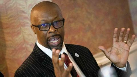 A man with a shaved head is wearing black framed glasses, a black pinstripe suit, white shirt and red tie. He has his arms near his face as he gestures