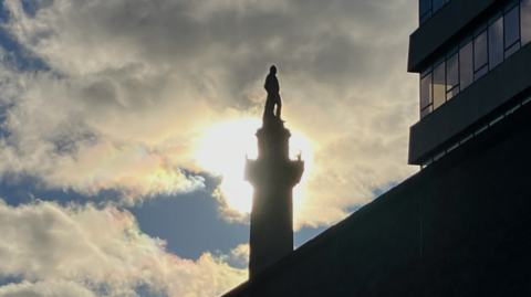The silhouette of a statue of a man on a tall plinth is set against a cloudy blue sky and a building in the foreground.