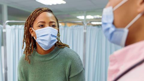 A woman wearing casual clothing sits on a hospital bed and speaks with her doctor. Both are wearing blue face masks.