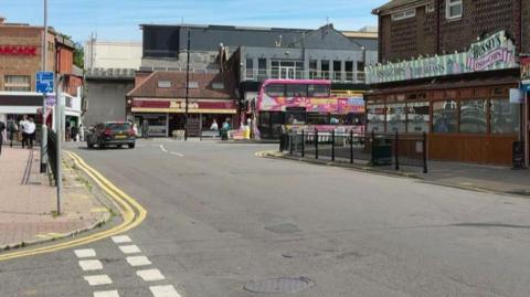 A view of Drummond road.  There is fish and chip shop in the building to the right, and a pink double decker bus crosses the t junction at the bottom of the road.