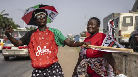 Two supporters of John Mahama pose by the roadside for a photograph in Accra on 3 December 2024. One is wearing the emblem of the National Democratic Congress - an umbrella - as a hat