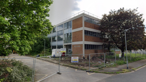 A Google street view screenshot of a brick building in an industrial site with trees on either side. The site is fenced off.