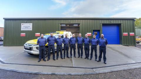 Nine men wearing navy trousers and blue shirts stand in a crescent-shape in front of a green building with an emergency vehicle parked outside.