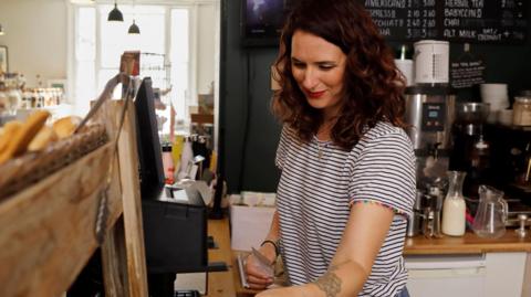 woman with shoulder-length brown hair in stripey t-shirt cleaning tables in a cafe