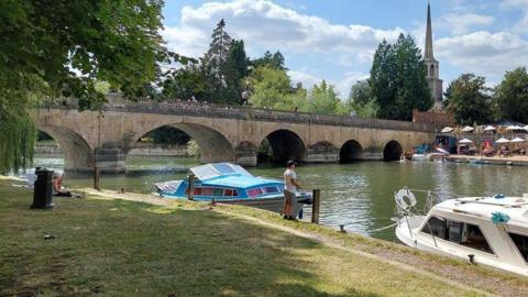 Wallingford riverside on a sunny day. A man is standing by the water while other residents are sitting near the bridge. There are more people walking on the bridge. There are two boats in the water. Across the river, people are sitting in an outdoor area under umbrellas.