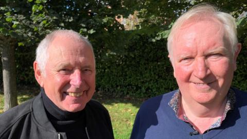 Nick Owen and Ian Phillipson standing together and smiling. Nick is wearing a black jacket and black turtle-neck jumper. Ian is wearing a blue jumper over a colourful shirt.