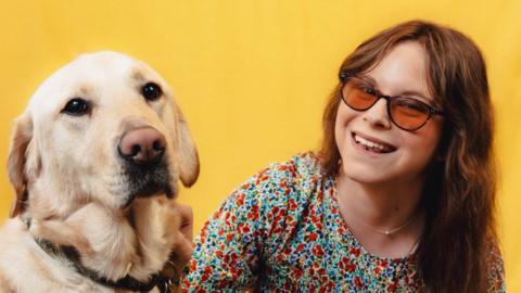 A photography studio image of a blonde guide dog looking at the camera, with Chloe his owner on the right. She is wearing sunglasses and a flowery dress. The background is yellow. 