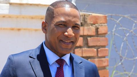 Duma Boko in a dark blue suit with red and blue stripped tie smiles outside his home in Botswana's capital, Gaborone. Behind him is a brick wall and barbed wire - November 2024