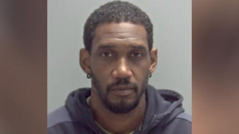 Tyrone Busch has short black hair in his custody photo and is looking at the camera. He is wearing a green top and a dark grey or navy blue hoodie over the top of it.