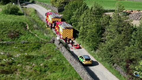 two fire engines and an emergency response vehicle are parked on a country road with fields and tall trees on either side