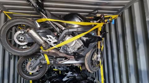 Multiple dark-coloured motorbikes packed into a container, held and elevated with yellow straps.