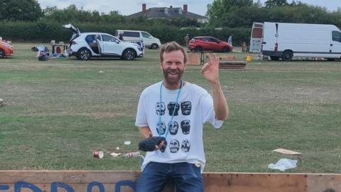 Paul Blakemore, a man with a beard, is carrying a microphone and perching on a wooden sign in a field.