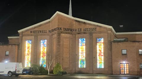 The church is a brown stone building with a white spire coming out of its roof. There are four large stained glass windows. And a car park in front of the building.