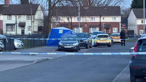 A residential street that is cordoned off with police tape. There are two police cars in the background and an officer standing, with their back to the camera, near the tape. There are houses and other cars surrounding the scene.