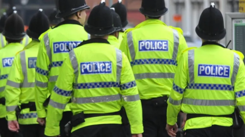 A group of police officers walking away from the camera. The officers are wearing black helmets and yellow high-vis jackets with 'police' written on the back